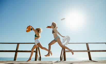 Seitenansicht von zwei Frauen, die auf einer Strandpromenade laufen und sich ihrer Kleidung entledigen. Freundinnen, die in ihrem Sommerurlaub im Meer schwimmen gehen. - JLPSF13409