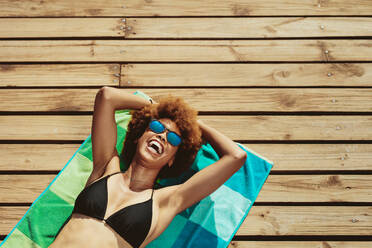 Draufsicht auf eine junge afrikanische Frau, die auf der Strandpromenade liegt und lächelt. Frau im Bikini entspannt sich am Strand. - JLPSF13412