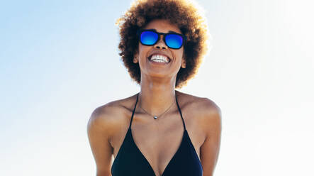 Attraktive junge Frau in Bikini und Sonnenbrille schaut weg und lächelt am Strand. Glückliches afrikanisches weibliches Modell gegen hellen Himmel im Freien. - JLPSF13419