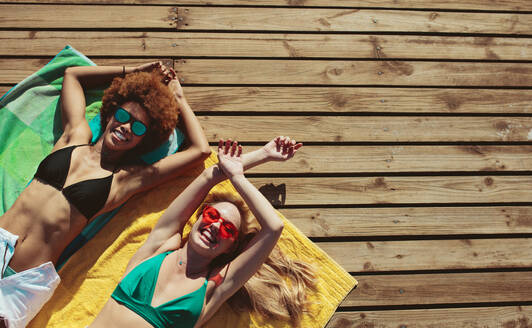 Draufsicht auf zwei junge Freundinnen, die auf einer Holzpromenade liegen. Zwei Frauen entspannen sich in ihrem Urlaub am Strand. - JLPSF13422