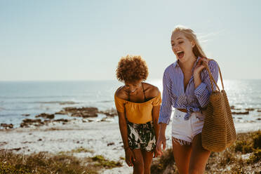 Zwei glückliche Frauen, die am Strand spazieren gehen. Lächelnde Freundinnen beim gemeinsamen Spaziergang am Meer. - JLPSF13426