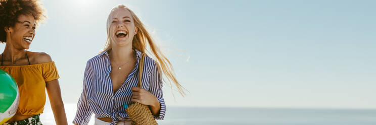 Beste Freundinnen im Urlaub an einem sonnigen Tag. Zwei junge Frauen, die zusammen am Strand spazieren gehen und lächeln, mit viel Platz zum Kopieren. - JLPSF13429