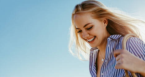 Lächelnde junge Frau vor blauem Himmel. Schöne kaukasische Frau mit hellem Sonnenlicht von hinten. - JLPSF13430