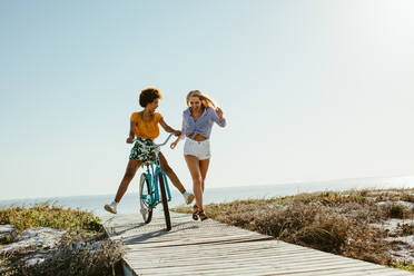 Zwei junge Frauen, die sich mit einem Fahrrad am Strand vergnügen. Frau, die mit einem Freund auf einem Fahrrad auf der Strandpromenade läuft. - JLPSF13431