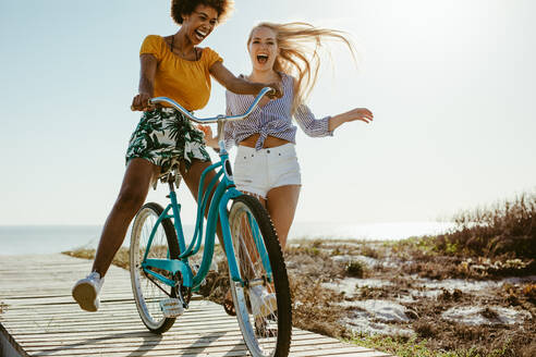 Fröhliche Freundinnen haben Spaß mit einem Fahrrad im Freien. Zwei Mädchen spielen mit einem Fahrrad an der Strandpromenade am Meer. - JLPSF13432