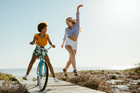 Afrikanisches Mädchen radelt auf einer Promenade mit ihren Freundinnen, die vor Begeisterung rennen und springen. Zwei Freundinnen, die sich im Urlaub amüsieren. - JLPSF13433
