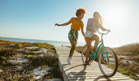 Eine aufgeregte Frau fährt mit dem Fahrrad die Strandpromenade entlang, während ihre Freunde rennen. Zwei junge Freundinnen haben eine tolle Zeit in ihrem Urlaub. - JLPSF13434