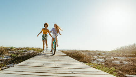 Zwei befreundete Frauen mit einem Fahrrad auf der Strandpromenade. Die Frau fährt mit dem Fahrrad, ihre Freunde halten sich von hinten fest und laufen. - JLPSF13435