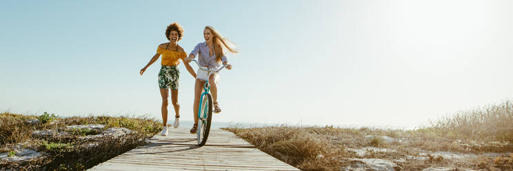 Aufgeregte Frau auf dem Fahrrad entlang der Promenade mit ihren Freundinnen, die mitlaufen. Zwei Freundinnen, die eine tolle Zeit in ihrem Urlaub haben. Panoramaaufnahme mit viel Kopierraum im Hintergrund. - JLPSF13436