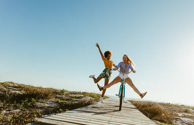 Excited woman riding bike down the boardwalk with her friends running behind. Two young female friends having a great time on their vacation. - JLPSF13437