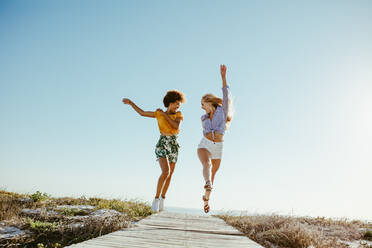 Two excited girls running and jumping with joy on boardwalk along the seaside. Two women friends having fun on their vacation. - JLPSF13438