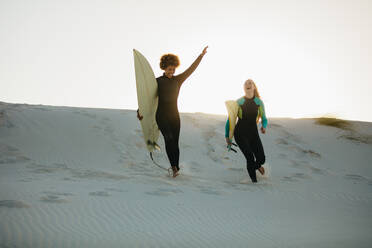 Aufgeregte junge Surferinnen, die den Strand entlang zum Meer laufen. Zwei Mädchen, die bei Sonnenuntergang mit Surfbrettern ins Meer laufen. - JLPSF13442