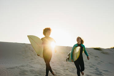 Zwei Surferinnen laufen bei Sonnenuntergang mit langen Surfbrettern auf das Meer zu. Glückliche junge Frauen, die im Meer surfen. - JLPSF13444