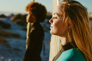 Nahaufnahme einer jungen kaukasischen Frau, die ein Surfbrett in der Hand hält, mit ihrer Freundin im Hintergrund. Zwei Surferinnen am Strand bei Sonnenuntergang. - JLPSF13446