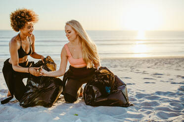 Two women picking up trash from the beach. Females cleaning the beach and collecting the litter in garbage bag. - JLPSF13456