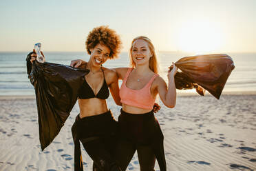 Zwei junge weibliche Freiwillige mit Müllsack. Zwei Surfer, die mit Plastikmüllsäcken am Strand stehen, mit Sonnenuntergang im Hintergrund. - JLPSF13459