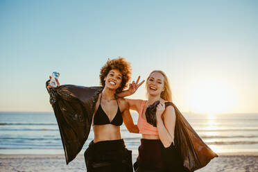 Cheerful female volunteers with garbage bag gesturing victory sign. Woman surfers on the beach with garbage bags during sunset. - JLPSF13460