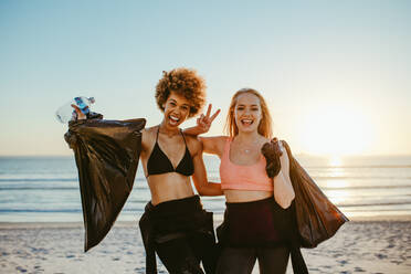 Two female surfers after picking up litter on the beach. Female volunteers after cleaning the beach area, standing together smiling and gesturing victory hand sign. - JLPSF13461