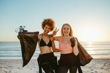 Zwei Surferinnen nach dem Aufsammeln von Müll am Strand. Freiwillige Frauen nach der Reinigung des Strandes, die lächelnd beieinander stehen und das Handzeichen für den Sieg geben. - JLPSF13461