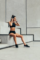 Fitness woman in sports clothing sitting on steps with headphones looking away. Female athlete taking rest after physical training. - JLPSF13479
