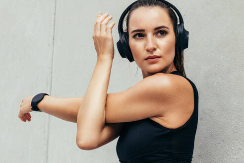 Fit young woman stretching her arms outdoors. Woman in sportswear and heaphones doing warmup exercise in the morning against grey wall. - JLPSF13484