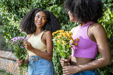 Happy female friends holding flowers in front of plant - DLTSF03222