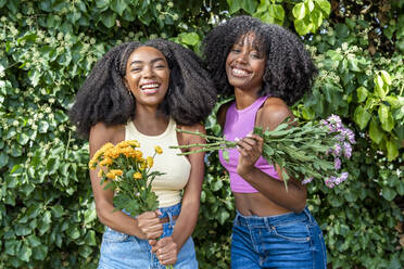 Happy friends holding flowers in front of plant - DLTSF03224