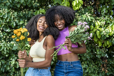 Smiling female friends holding flowers in front of plant - DLTSF03225