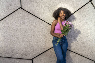 Glückliche Frau hält lila Blumen vor einer Mauer - DLTSF03235