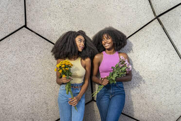 Happy young woman with friend holding flowers leaning on wall - DLTSF03237