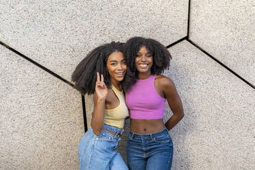 Smiling woman gesturing peace sign standing with friend in front of wall - DLTSF03244