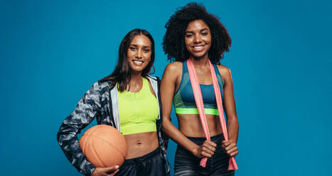 Zwei junge Frauen stehen zusammen mit einem Basketball und einem Widerstandsband vor einem blauen Hintergrund. Gemischtrassige Freundinnen in Sportkleidung schauen in die Kamera und lächeln. - JLPSF13560