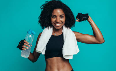 Junge Fitness-Frau, die ihre Muskeln anspannt und lächelt. Afrikanisches weibliches Model in Sportkleidung, das eine Wasserflasche und ein Handtuch um den Hals hält und ihre Muskeln zeigt. - JLPSF13589