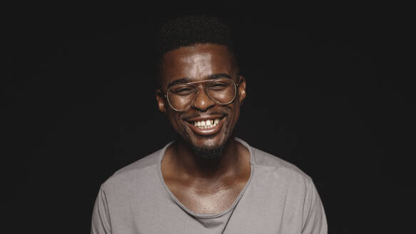 Lachender afrikanischer Mann mit Brille isoliert auf schwarzem Hintergrund. Nahaufnahme eines glücklichen afrikanischen Mannes, der in die Kamera schaut. - JLPSF13635