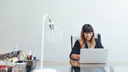 Lächelnde kreative Künstlerin sitzt an ihrem Schreibtisch im Büro und arbeitet an einem Laptop. Illustratorin sitzt im Büro und arbeitet an einem Laptop. - JLPSF13662
