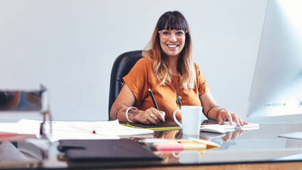 Illustratorin, die eine Skizze auf einem digitalen Schreibblock anfertigt. Lächelnde Unternehmerin, die an ihren Entwürfen arbeitet, während sie an ihrem Schreibtisch im Büro sitzt. - JLPSF13676