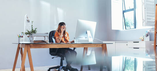 Fröhliche Frau, die über ein Mobiltelefon spricht und auf einen Computer im Büro schaut. Lächelnde Geschäftsfrau, die an ihrem Tisch im Büro sitzt und an einem Computer arbeitet. - JLPSF13685