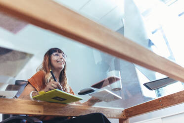 Low angle view of a female illustrator making a drawing on a digital pad looking at the computer. Smiling creative artist working on her designs on a computer placed on a transparent glass top table. - JLPSF13686