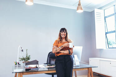 Lächelnde Unternehmerin, die in der Nähe ihres Tisches im Büro steht und eine Kaffeetasse hält. Fröhliche Geschäftsfrau in ihrem Büro, die eine Tasse Kaffee genießt. - JLPSF13688