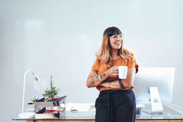 Porträt einer lächelnden Geschäftsfrau, die an ihrem Schreibtisch steht, eine Kaffeetasse hält und wegschaut. Eine Unternehmerin, die eine Pause macht und eine Tasse Kaffee genießt. - JLPSF13689