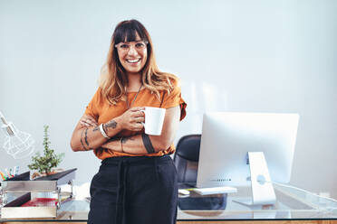 Lächelnde Geschäftsfrau mit einer Kaffeetasse in der Hand. Porträt einer kreativen Unternehmerin, die neben ihrem Schreibtisch steht und eine Tasse Kaffee genießt. - JLPSF13690