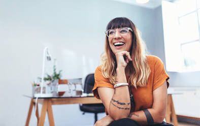 Nahaufnahme einer Geschäftsfrau, die lachend im Büro sitzt und ihr Kinn auf die Hand stützt. Lächelnde Unternehmerin mit Brille, die eine Pause von der Arbeit macht. - JLPSF13695