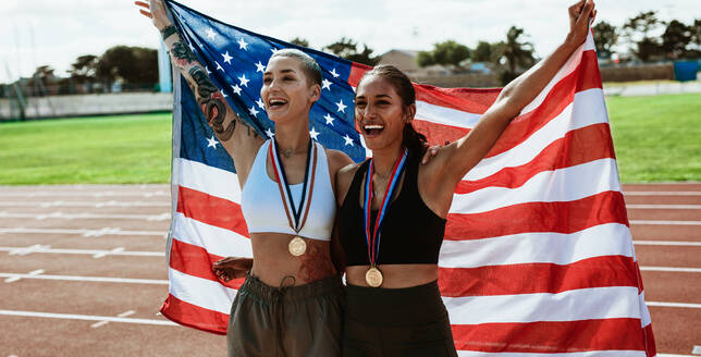 Weibliche Athleten auf der Laufbahn feiern den Sieg und halten die amerikanische Flagge. Der Sprinter trägt die US-Flagge und schreit nach dem Sieg. - JLPSF13704