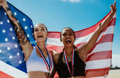 Weibliche Athleten feiern ihren Sieg mit der amerikanischen Flagge im Stadion. Jubelnde Läuferinnen, die nach ihrem Sieg die US-Flagge halten. - JLPSF13705