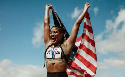 Side view of confident young ethnic lady in sports bra waving American flag  and looking at camera against modern glass skyscraper stock photo