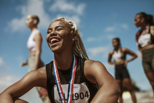 Lächelnde Sportlerin nach gewonnenem Rennen mit anderen Teilnehmern im Hintergrund. Sportlerin mit Medaille, die ihren Sieg im Stadion feiert. - JLPSF13727