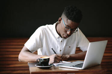 Freelancer making notes sitting at a coffee table. Man working on his laptop computer at a coffee shop. - JLPSF13764