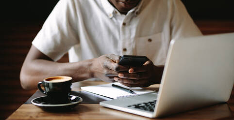 Ein Mann benutzt sein Handy, während er in einem Café an seinem Laptop arbeitet. Ausschnitt eines Freiberuflers, der mit seinem Laptop, seinem Handy und seinen Notizen an einem Kaffeetisch sitzt. - JLPSF13768