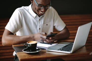 Ein Mann schaut auf sein Handy, während er in einem Café an seinem Laptop arbeitet. Ein Freiberufler, der mit seinem Laptop, seinem Handy und seinen Notizen an einem Kaffeetisch sitzt und seine Arbeit erledigt. - JLPSF13769