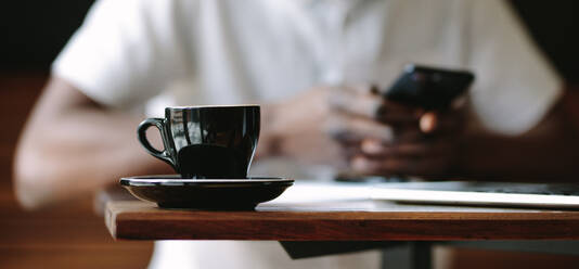 Closeup of a coffee cup placed on a table in a coffee shop. Cropped shot of man sitting at a coffee shop with a glossy black coffee cup and saucer on the table. - JLPSF13770